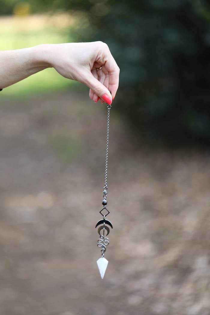 A detailed view of a hand holding a crystal pendulum outdoors with a blurred natural background.